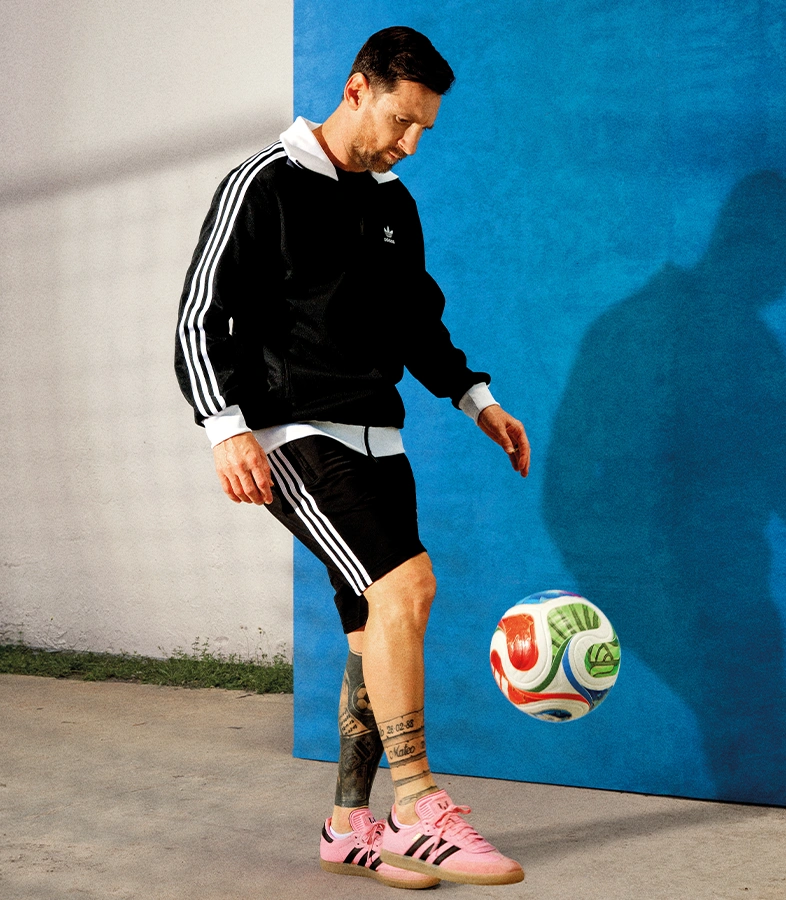 Lionel Messi wearing a black adidas track top and shorts balances a football in front of a blue and white background while wearing pink adidas sneakers. (Photo)