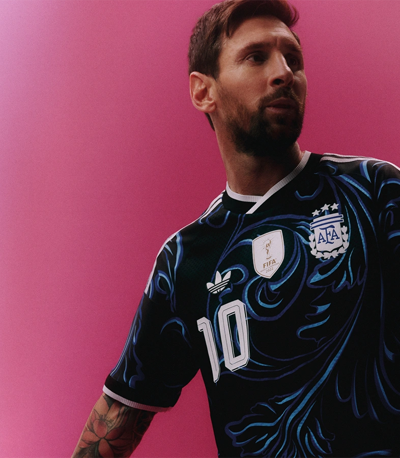 Lionel Messi wearing a black adidas track top and shorts balances a football in front of a blue and white background while wearing pink adidas sneakers. (Photo)