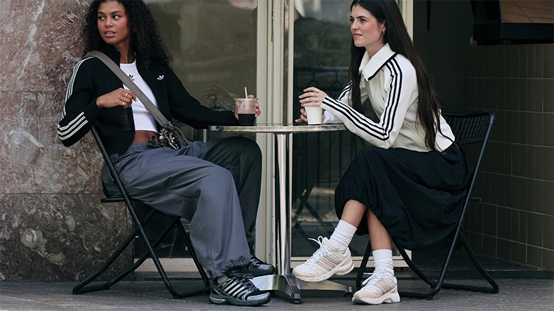 Two visible women sitting at a small outdoor café table, wearing sporty outfits with three stripes and holding drinks. (Photo)