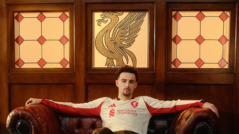 Man wearing a white and red Liverpool jersey sitting with arms outstretched on a leather sofa in front of wood paneling with the club crest. (Photo)