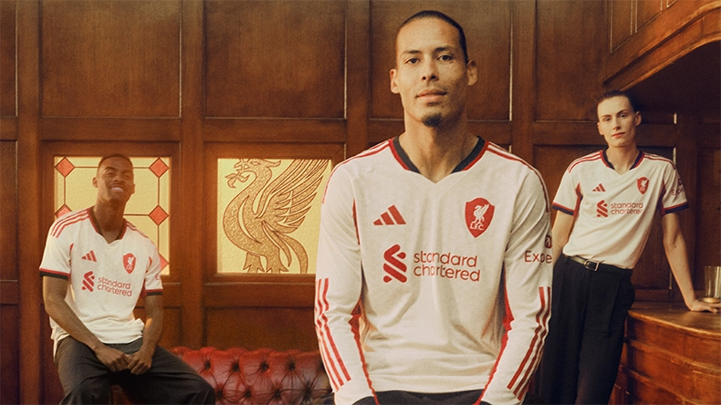 Three men in white Liverpool jerseys standing and sitting in a wood-paneled room with the club crest in the background. (Photo)