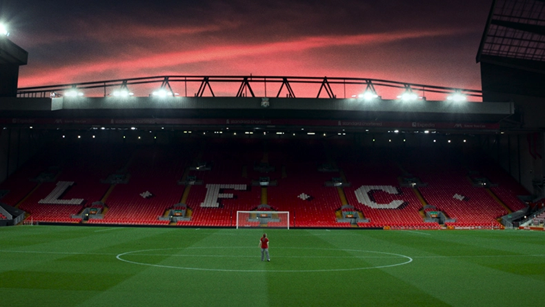 Wide-angle view of a football stadium at sunset with red seats spelling “L.F.C.”; a single person stands on the pitch. (Photo)